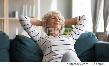 Smiling older woman leaning back, relaxing on cozy couch 64320089