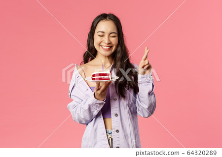 Celebration, holidays and people concept. Portrait of cheerful carefree, pretty asian girl cross fingers, close eyes and smiling joyfully while making wish before blowing candle on birthday cake 64320289