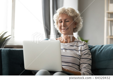 Smiling older woman looking at laptop screen, sitting on couch 64320432