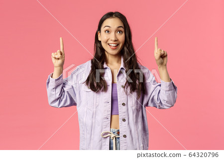 Enthusiastic young pretty girl looking and pointing fingers up with happy smile, see promo, interested in taking part event, party, found great online courses, standing pink background 64320796