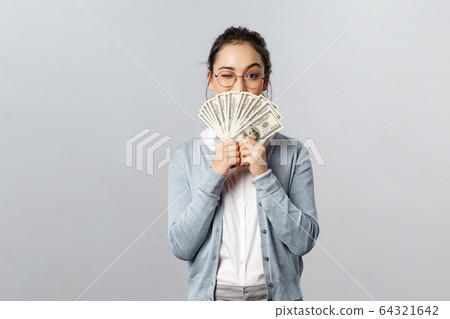 Business, finance and investment concept. Happy, pleased pretty asian woman, rich girl playfully raise eyebrow hiding satisfied smile behind money dollars, win cash prize, grey background 64321642