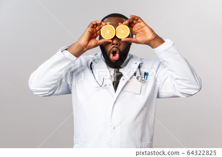 Healthcare, healthy diet and disease concept. Funny good-looking physician, african-american doctor playing with orange, look amused, tell stay safe, eat vitamins and fruits, grey background 64322825