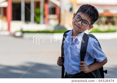 A cute Asian primary school boy wearing glasses and a white school uniform, carrying a black backpack, standing in front of the school. A cute Asian primary school boy wearing glasses and a white school uniform, carrying a black backpack, standing in front of the school. 64326474