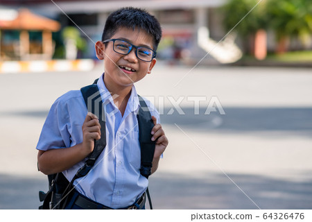 A cute Asian primary school boy wearing glasses and a white school uniform, carrying a black backpack, standing in front of the school. A cute Asian primary school boy wearing glasses and a white school uniform, carrying a black backpack, standing in front of the school. 64326476