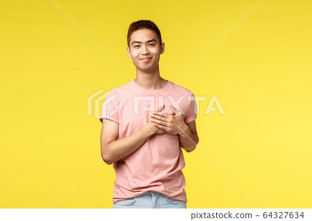 People, different emotions and lifestyle concept. Portrait of touched asian guy holding hands on heart and smiling pleased, keep good memories in soul, being grateful for help, yellow background 64327634