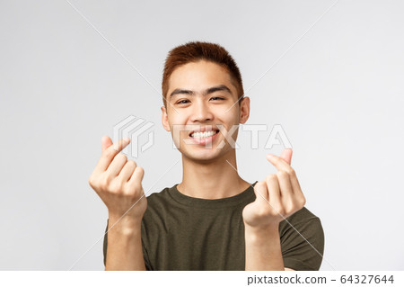 People, different expressions and lifestyle concept. Cute asian guy with stylish haircut, showing korean finger hearts and smiling lovely, express sympathy or like, stand grey background People, different expressions and lifestyle concept. Cute asian guy with stylish haircut, showing korean finger hearts and smiling lovely, express sympathy or like, stand grey background 64327644