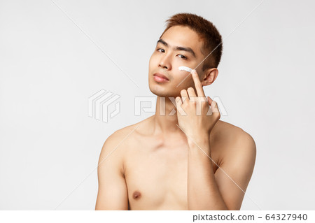 Beauty, people and leisure concept. Sassy good-looking asian man with naked torso taking care of his perfect, no blemishes skin, apply cream on face and look camera pleased, white background 64327940