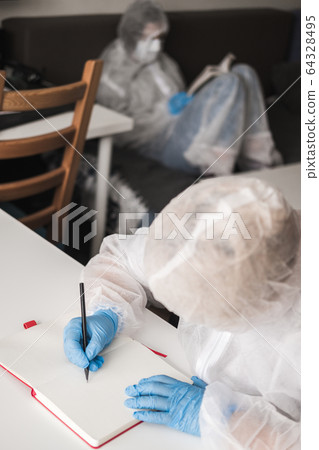 Girl in protective white translucent suit, blue rubber gloves, medical mask sits at table at home and draws in sketchbook with pencil. Remote work of designer, artist during coronavirus pandemic. Girl in protective white translucent suit, blue rubber gloves, medical mask sits at table at home and draws in sketchbook with pencil. Remote work of designer, artist during coronavirus pandemic. 64328495