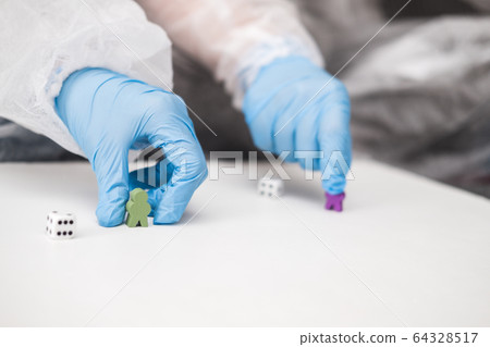 Two people in protective white translucent suit, blue rubber gloves, medical mask sit on sofa at home and play board games, roll dice. Stay home during coronavirus pandemic covid. Two people in protective white translucent suit, blue rubber gloves, medical mask sit on sofa at home and play board games, roll dice. Stay home during coronavirus pandemic covid. 64328517