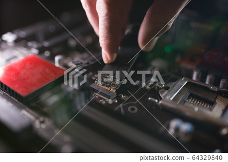 Focus at SSD slot, type M.2 with support for NVMe, on computer motherboard. Technician is installing new fast high capacity drive with flash chips, known as SSD. 64329840