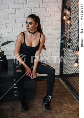 Beautiful fashionable woman at loft style room with white bricks wall and black iron net. Attractive girl with perfect figure in stylish black clothes. Beautiful fashionable woman at loft style room with white bricks wall and black iron net. Attractive girl with perfect figure in stylish black clothes. 64330341