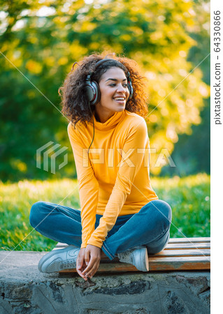 African teenager girl listens to music with headphones in park. Cute woman in yellow, dancing with 64330866
