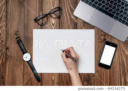 Woman's hand writes a pen to a sheet of white paper. Wooden office desk table with laptop, smart phone, clear white paper and office supplies. 64334273