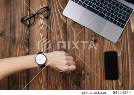 Woman's hand with watch. Top view business office supplies. Laptop, smart phone, glasses and watch on wooden table. Time to work. 64334274