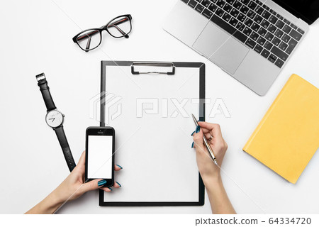 A woman's hand holds a pen and writes on a clean sheet of paper. White office desk with laptop, smartphone and supplies. A woman's hand holds a pen and writes on a clean sheet of paper. White office desk with laptop, smartphone and supplies. 64334720