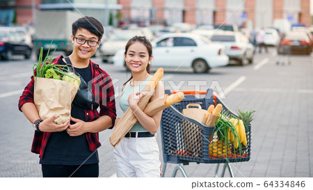 Likable asian couple hold paper eco bags with organic healthy food in hands while standing near store mall. Happy family shopping on weekend. Likable asian couple hold paper eco bags with organic healthy food in hands while standing near store mall. Happy family shopping on weekend. 64334846