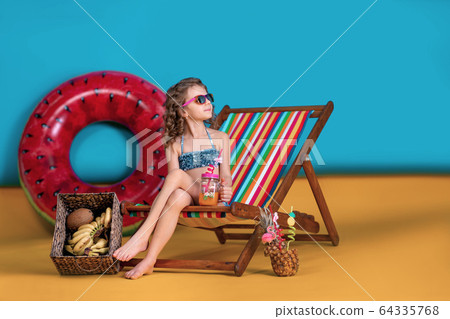 Girl wearing swimsuit, sunglasses holding jar with juice or cocktail with multicolored straws sitting in rainbow deck chair by big watermelon inflatable circle and looking away 64335768
