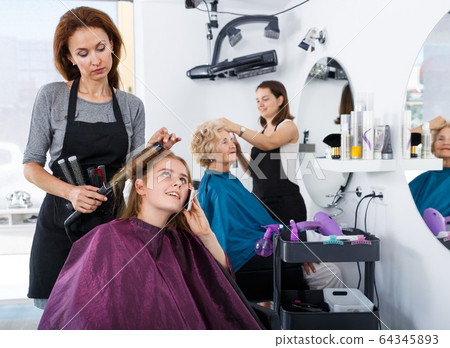 Stylist curling hair of teen girl with phone 64345893