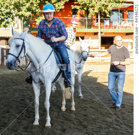 Man in helmet riding by horse at ranch, trainer talking to female Man in helmet riding by horse at ranch, trainer talking to female 64346171