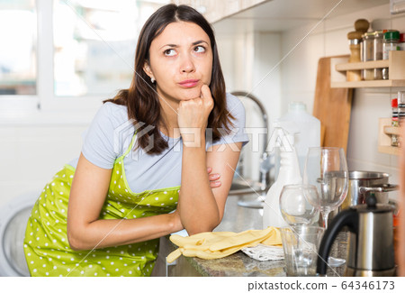 Tired girl housewife in apron holding gloves at kitchen Tired girl housewife in apron holding gloves at kitchen 64346173