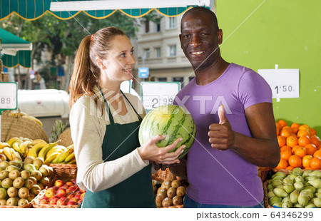 Smiling seller woman is helping man choose melone and watermelon Smiling seller woman is helping man choose melone and watermelon 64346299