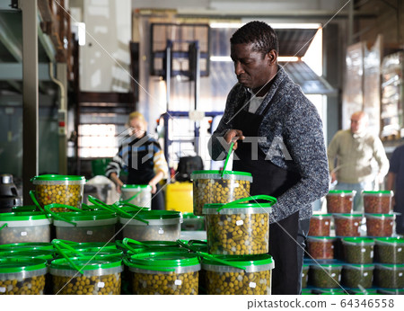 Afro-american male worker stocks plastic containers and cans with olives in warehouse Afro-american male worker stocks plastic containers and cans with olives in warehouse 64346558