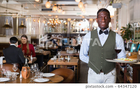 Man waiter is holding tray with order in hall of restaurant. Man waiter is holding tray with order in hall of restaurant. 64347120