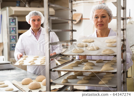 Female baker placing tray with formed dough on trolley 64347274