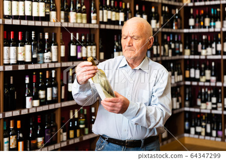 Confident grey-haired elderly man choosing white wine in modern wineshop 64347299