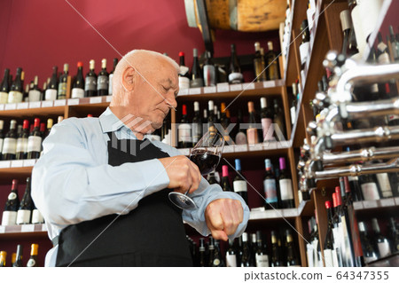 Confident elderly male winemaker holding glass of red wine, checking it in wine store Confident elderly male winemaker holding glass of red wine, checking it in wine store 64347355