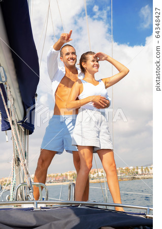 Loving couple standing on deck of sailboat during sea trip along Spain shoreline, looking and pointing to horizon 64348427