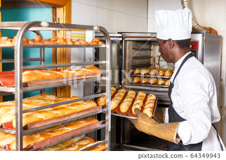 Worker of bakery putting baked baguettes on tray rack Worker of bakery putting baked baguettes on tray rack 64349943