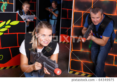Smiling young female holding laser gun during laser tag game with players 64349975