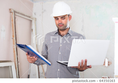 Professional constructor is standing with laptop and folder with documents Professional constructor is standing with laptop and folder with documents 64350163