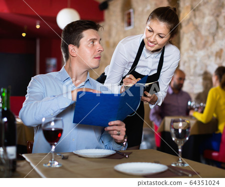 Hospitable waitress helping handsome man with menu, taking order in restaurant 64351024