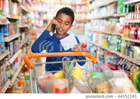Focused African tween boy talking on phone and looking at shopping list while visiting supermarket 64352243
