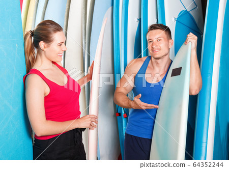 Young couple planning to surf, choosing boards and surfing suits in beach club Young couple planning to surf, choosing boards and surfing suits in beach club 64352244