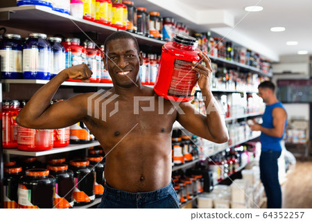 Bare-chested athletic African standing with cans of sports nutrition in store, demonstrating muscles Bare-chested athletic African standing with cans of sports nutrition in store, demonstrating muscles 64352257