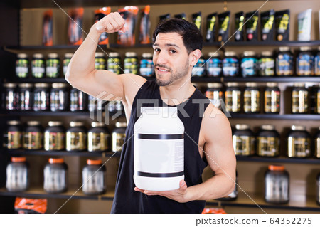 Young bodybuilder showing his biceps and holding pot of sport nutrition products in shop Young bodybuilder showing his biceps and holding pot of sport nutrition products in shop 64352275