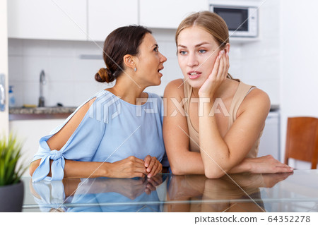Upset young women friends whispers in the ear and talking at table in home Upset young women friends whispers in the ear and talking at table in home 64352278