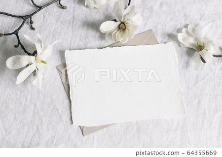 Wedding stationery mock-up scene. Blank horizontal greeting card, envelope on linen tablecloth background with white magnolia stellata tree branches and ribbon. Feminine still life. Flat lay, top view 64355669