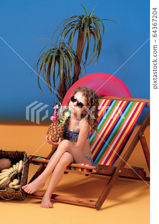 Smiling girl in swimsuit and sunglasses sitting in rainbow deck chair holding pineapple cocktail with colorful straws and showing thumb up and sunbathing Smiling girl in swimsuit and sunglasses sitting in rainbow deck chair holding pineapple cocktail with colorful straws and showing thumb up and sunbathing 64367204