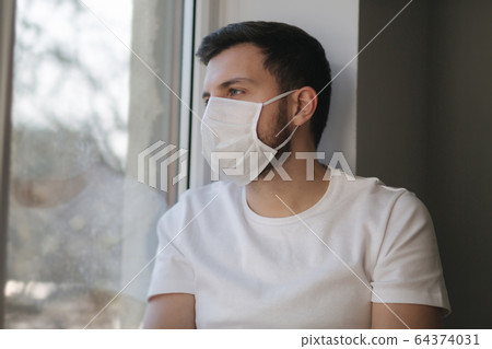 Young man sits on windowsill and out on protective mask and look s out the window. Alone at home in quarantine. Coronavirus theme. Stay at home Young man sits on windowsill and out on protective mask and look s out the window. Alone at home in quarantine. Coronavirus theme. Stay at home 64374031