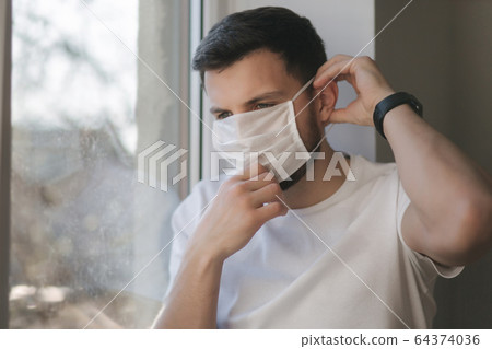Young man sits on windowsill and out on protective mask and look s out the window. Alone at home in quarantine. Coronavirus theme. Stay at home 64374036