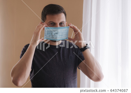 Man put on madical mask at home. Young man in protective mask. Coronavirus theme Man put on madical mask at home. Young man in protective mask. Coronavirus theme 64374087