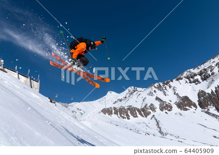 A male athlete skier in an orange trigger makes a jump jump with a grab with flying snow powder against the background of Mount Elbrus in the North Caucasus. Winter Extreme Sports Concept A male athlete skier in an orange trigger makes a jump jump with a grab with flying snow powder against the background of Mount Elbrus in the North Caucasus. Winter Extreme Sports Concept 64405909