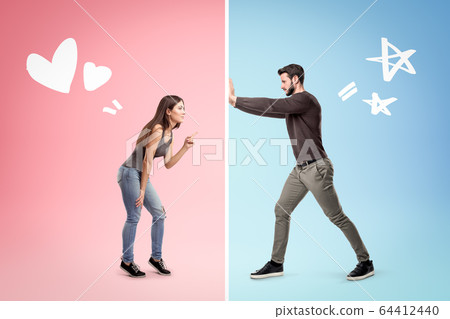 Side view of young girl and young man standing opposite each other on different backgrounds, pink and blue, man trying to break wall that separates them. 64412440