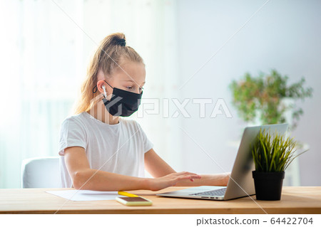 Serious schoolgirl sitting at table with laptop and textbook and doing homework. 64422704