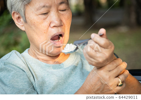Asian senior woman holding spoon and hands tremor while eating rice,cause of hands shaking include parkinson's disease,stroke, brain injury,symptom of essential tremor,health problem of elderly people 64429011