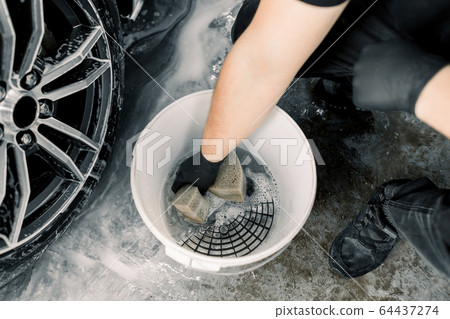 Car wash and detailing concept. Top view of washing tools in car wash service, white bucket with soap cleaning solution, special grille. Hand of male worker holding sponge for cleaning car rims 64437274